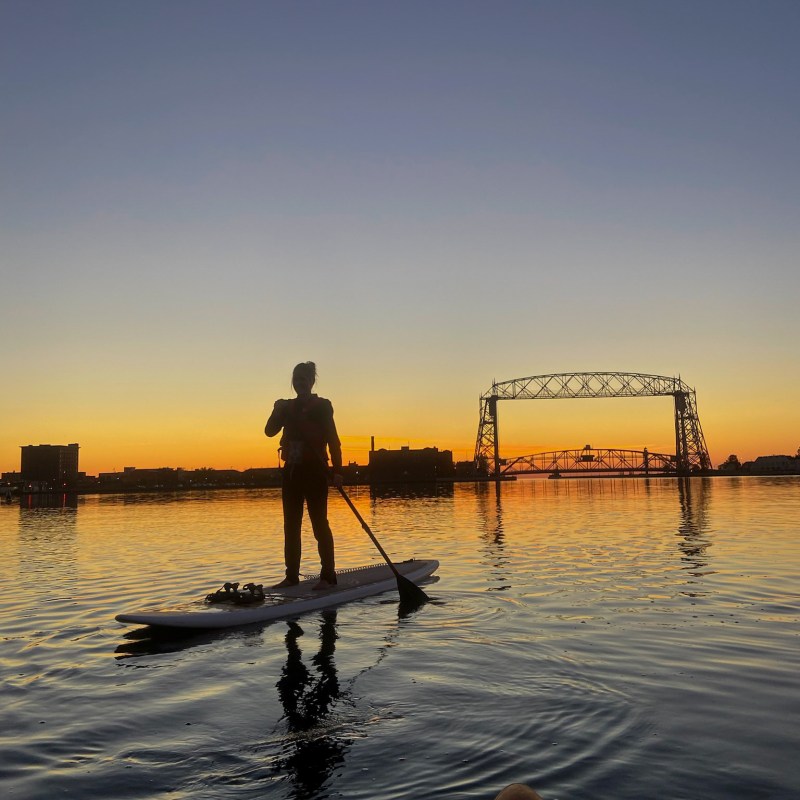 a woman on a paddle board in front of sunrise and Duluth bridge