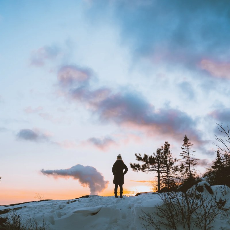 Silhouette of person on snowy hill at sunset, with trees and clouds.