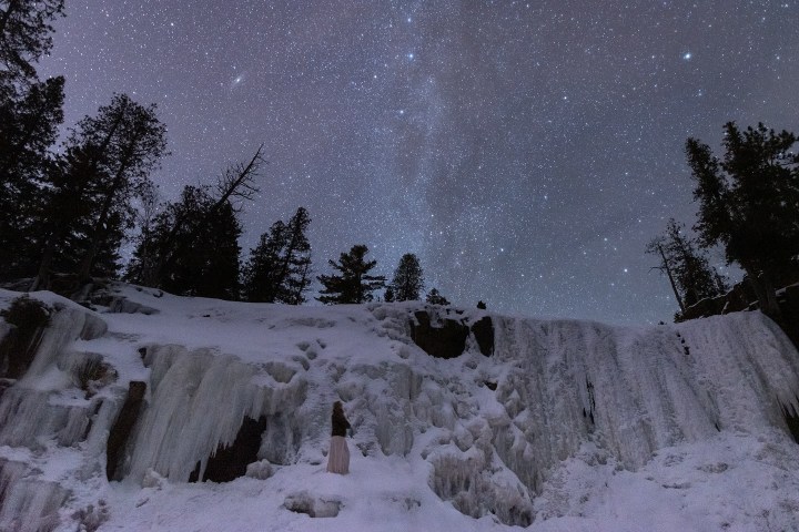 Starry night sky over snowy cliffs and silhouetted trees.