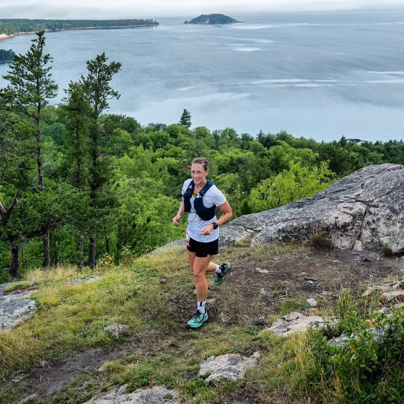 Person trail running on a rocky path with trees and a lake in the background.