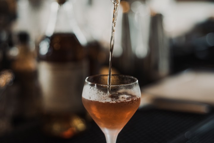 Hand pouring cocktail into a glass using a strainer at a bar.