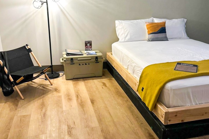 Minimalist bedroom with wood bed, yellow blanket, floor lamp, black chair, and storage box on wooden floor.