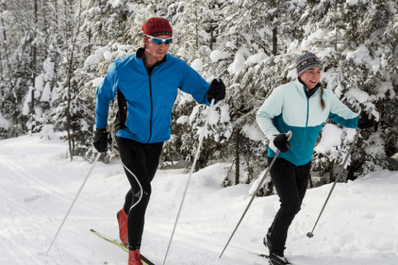 Two people cross-country skiing on a snowy trail surrounded by snow-covered trees.