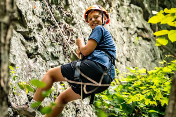 Person rock climbing with a harness and helmet in a forested area.