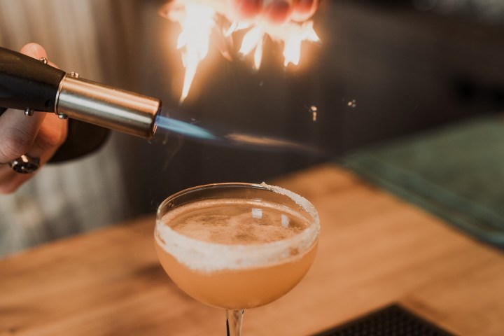 Bartender igniting citrus peel over a cocktail glass with a torch.
