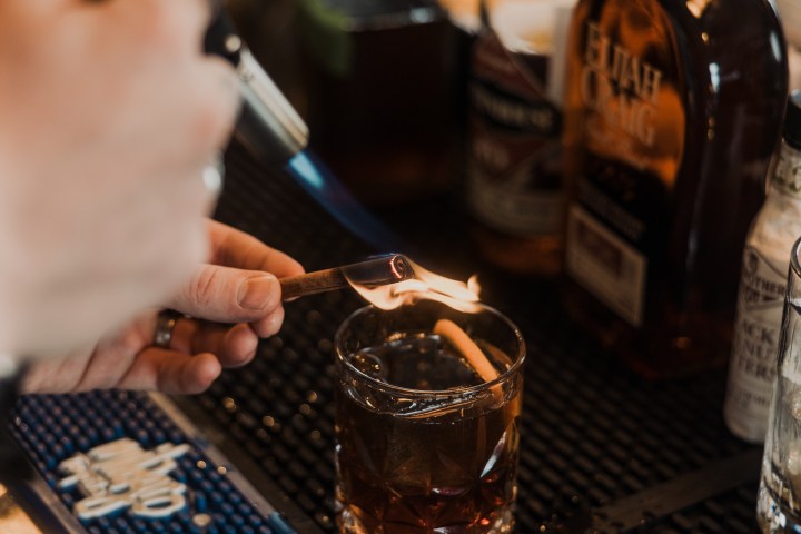 Bartender flaming an orange peel over a glass of cocktail with liquor bottles in the background.