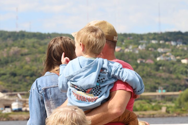 People standing on a boat deck, overlooking a river and hills.