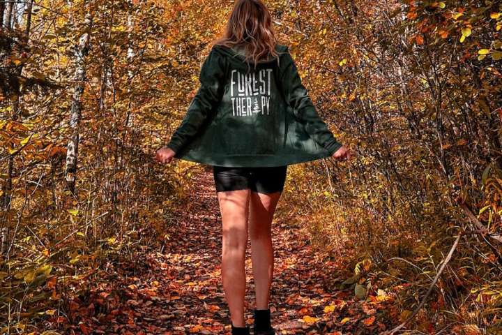 Person walking on a leafy forest path wearing a green hoodie, surrounded by autumn trees.