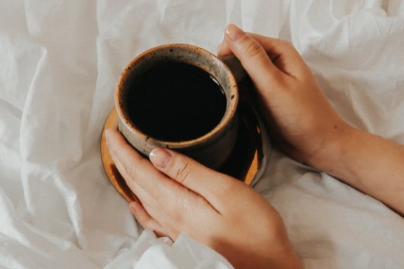 Hands holding a cup of coffee on a bed with white sheets.
