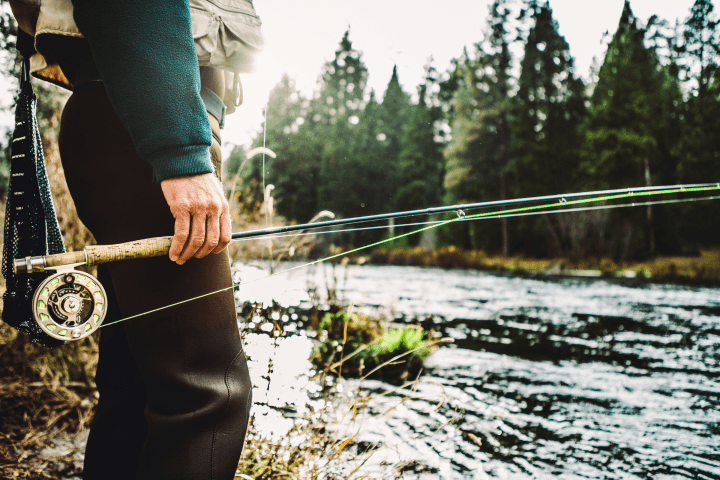 Person holding a fishing rod by a flowing river with trees in the background.
